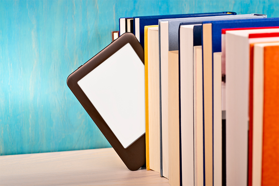 Una fila di libri appoggiati su un piano di legno, da cui fa capolino un e-reader