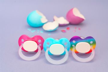 Tre ciucci colorati per neonatee neonati con i colori rosa, azzurro e arcobaleno. Sullo sfondo delle uova colorate rotte.