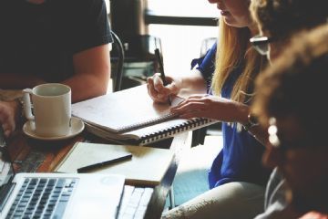 4 persone sedute ad una scrivania nella quale ci sono un pc e una persona che prende appunti.