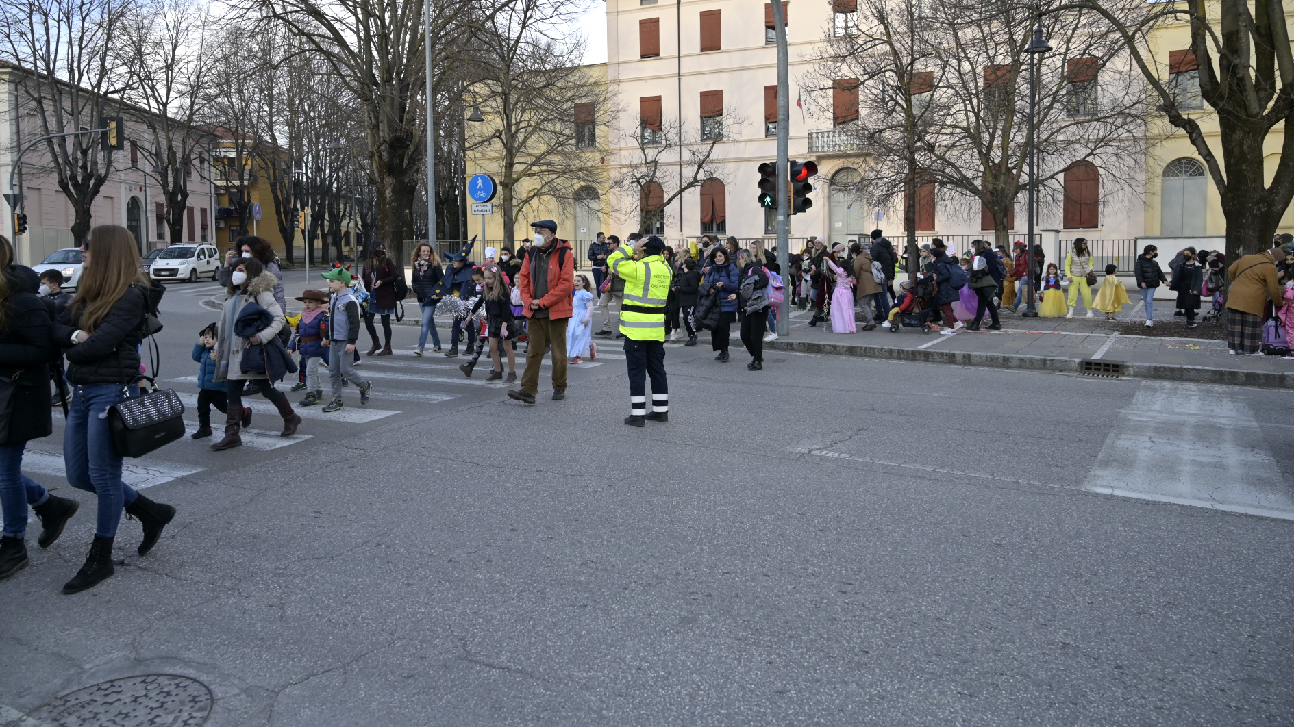Bambini e genitori che attraversano la strada con l'aiuto della polizia locale, in secondo piano la scuola.