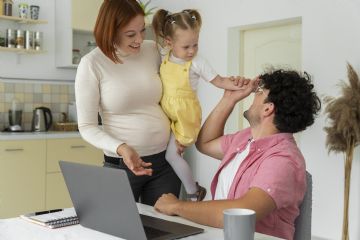 una coppia in cucina in cui l'uomo è seduto al tavolo di fronte al pc. La donna incinta ha in braccio anche una bambina piccola e interagiscono tutti e tre.